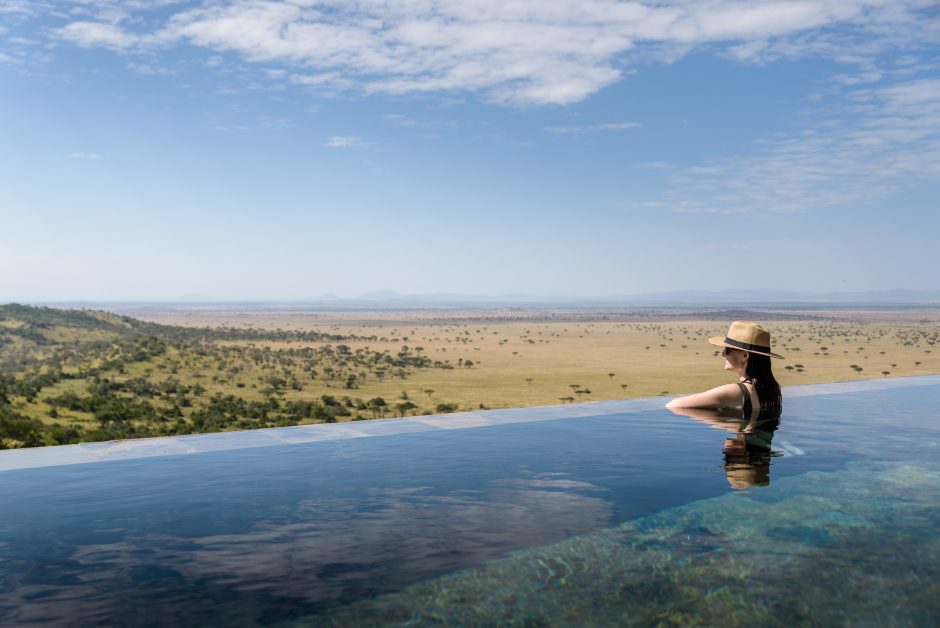 hillside_suite_guest_in_pool_overlooking_serengeti_adriaan_louw A woman in a straw hat relaxes in an infinity pool overlooking the vast Serengeti plains, with scattered acacia trees and distant hills under a clear blue sky.